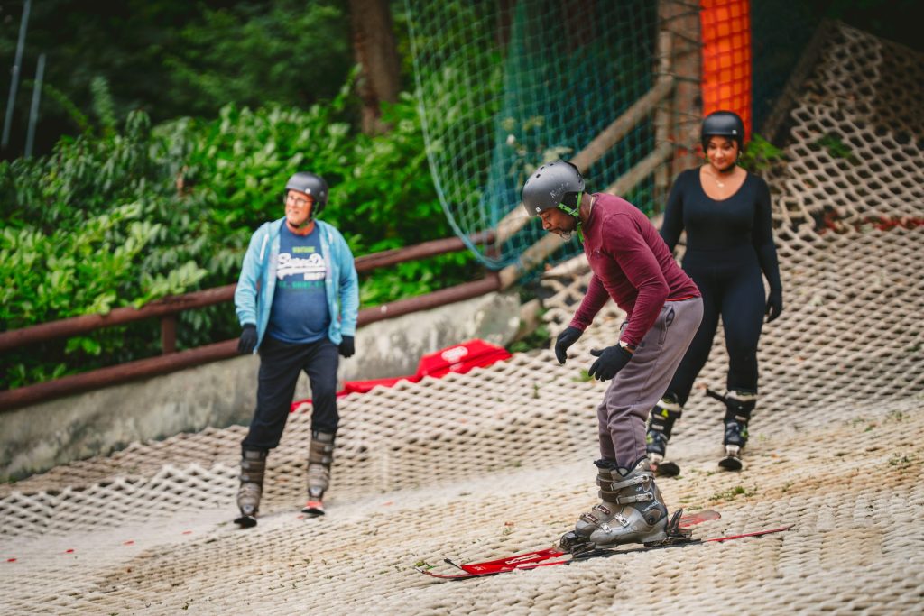 Two adults stand off to the side of a dry ski slope as another learner skis down during a lesson.