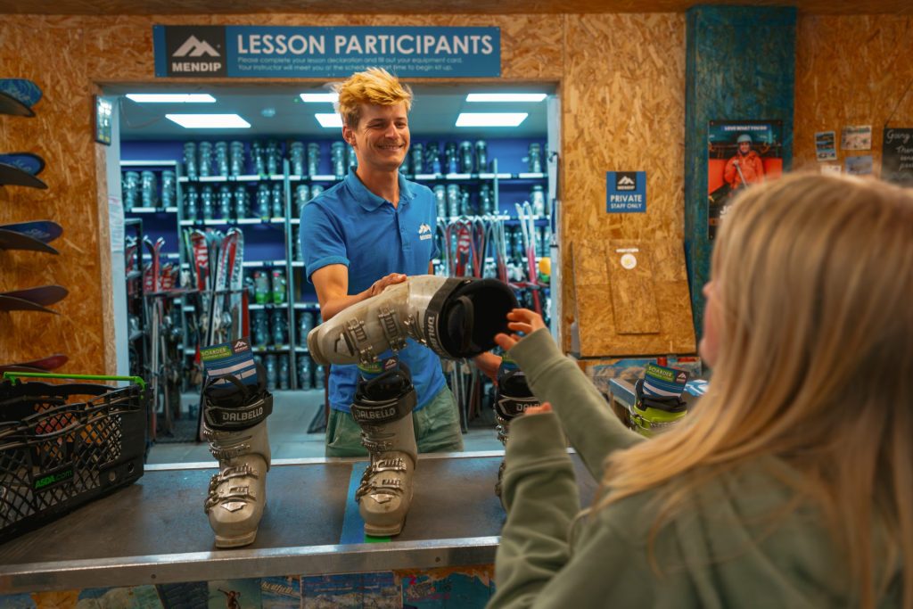 A member of staff is smiling as they hand a ski boot to a client. Ski and snowboard equipment is arranged on shelves behind them.
