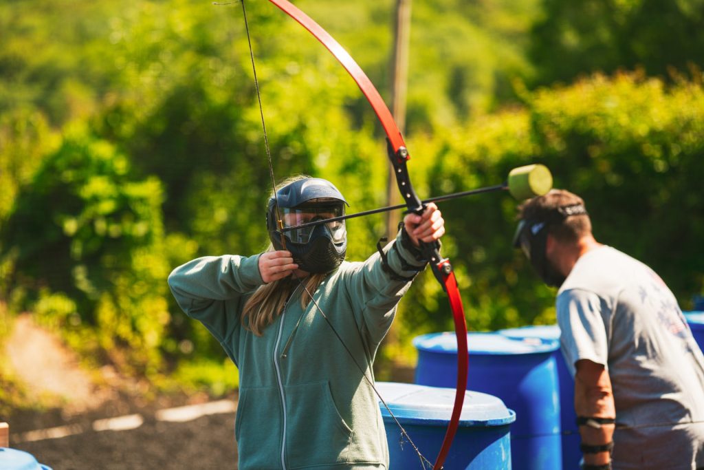 A woman wears a paintball-stye mask and green hoody. She is pulling back a foam-tipped arrow as she takes aim at the opposing team. This is one of the activities available to book when on a summer ski camp.