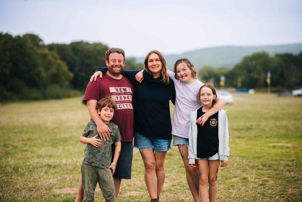 A family of five stands together with their arms around each others shoulders, smiling at the viewer. They are standing in a camping field decorated with flags that hosts a family festival with accommodation.