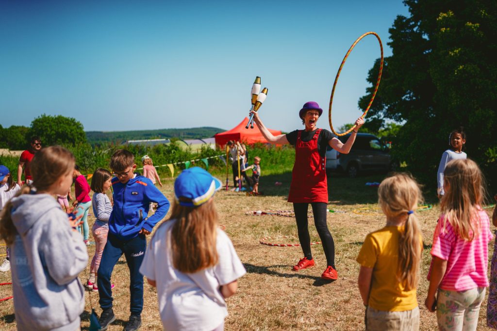 Children stand in a circle around a woman wearing a purple hat and red dress. They are watching her give a hoop demonstration at the Circus Skills activity. This is one of the free activities available during your summer ski camp.