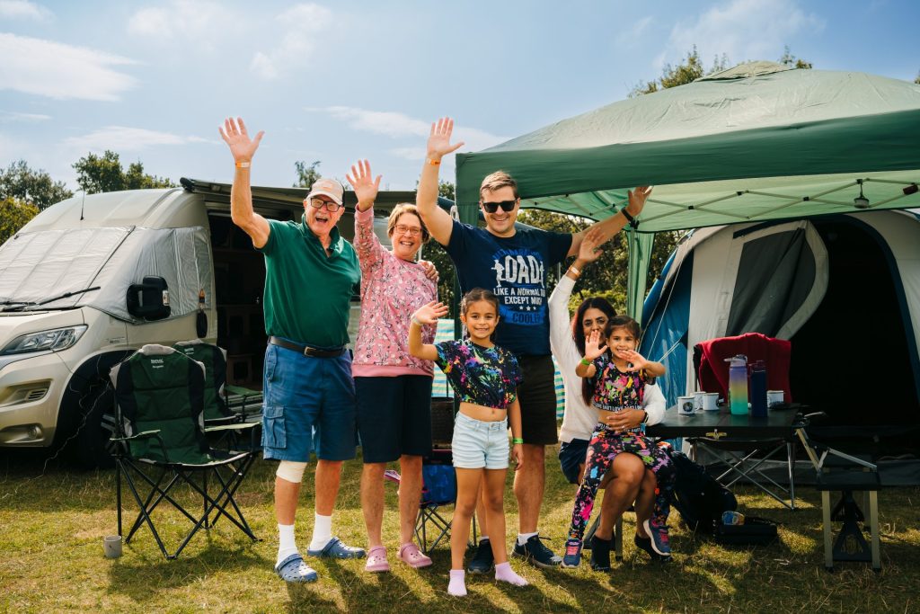 A mixed family group stands together next to their caravan and tent. They are all smiling and waving at the viewer, demonstrating the accommodation available for summer ski camp.