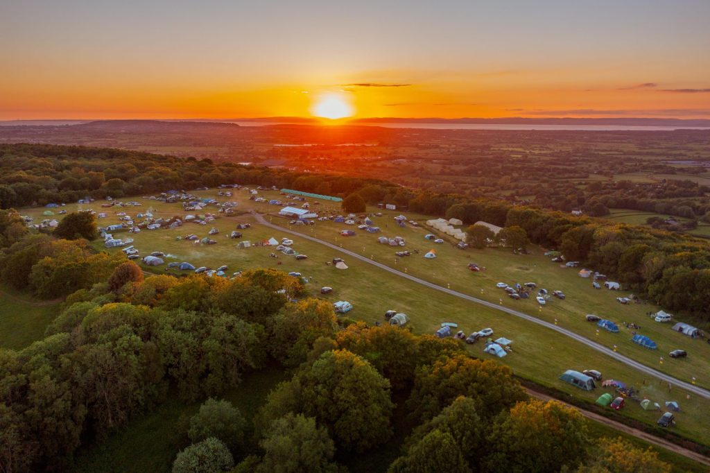 An aerial view of Basecamp, a campsite in the Mendip Hills perfect for a summer ski camp. It is a large field on top of a hill; the sun sets in the background and tents and caravans dot the camping field.