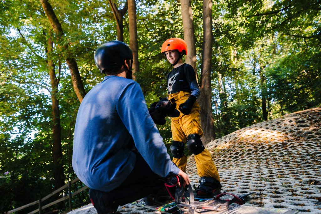 A child on a snowboard stands in front of an instructor, listening to them speak. They are on an all-weather dry slope available for summer ski camps.