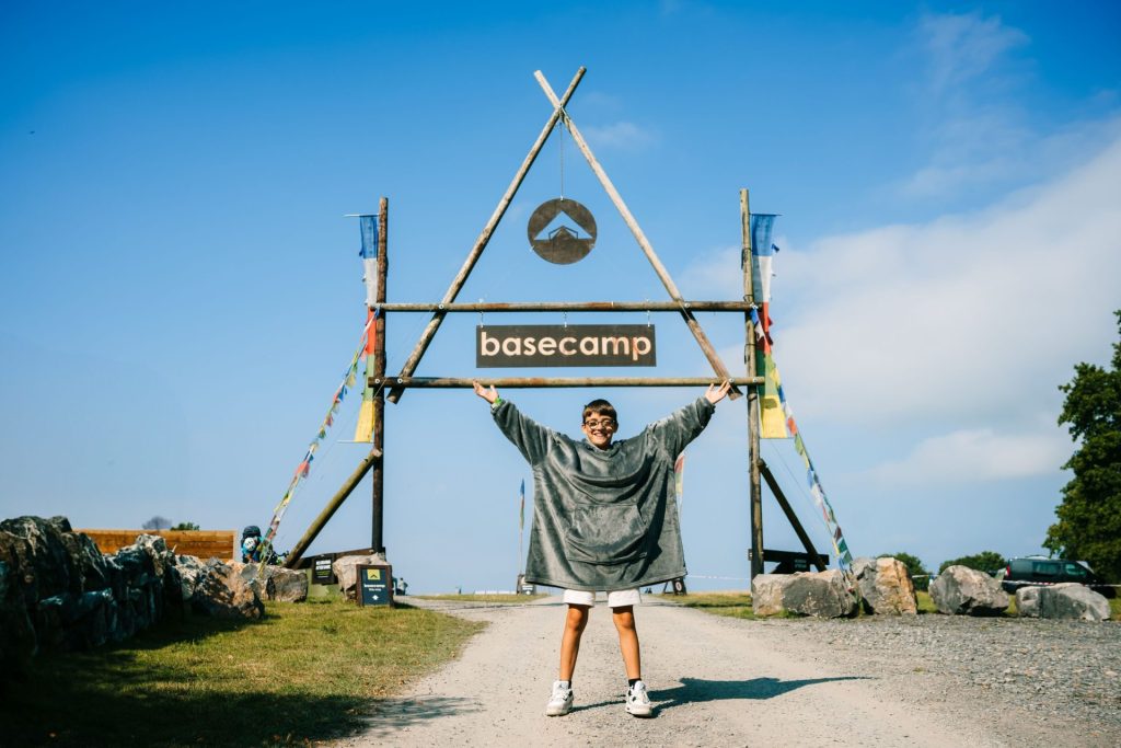 A boy stands in front of the entrance to Basecamp, a campsite near Bristol. He is smiling and holding his arms up in a welcoming way. The entry is a wooden archway decorated with flags and a sign that says 'Basecamp'.