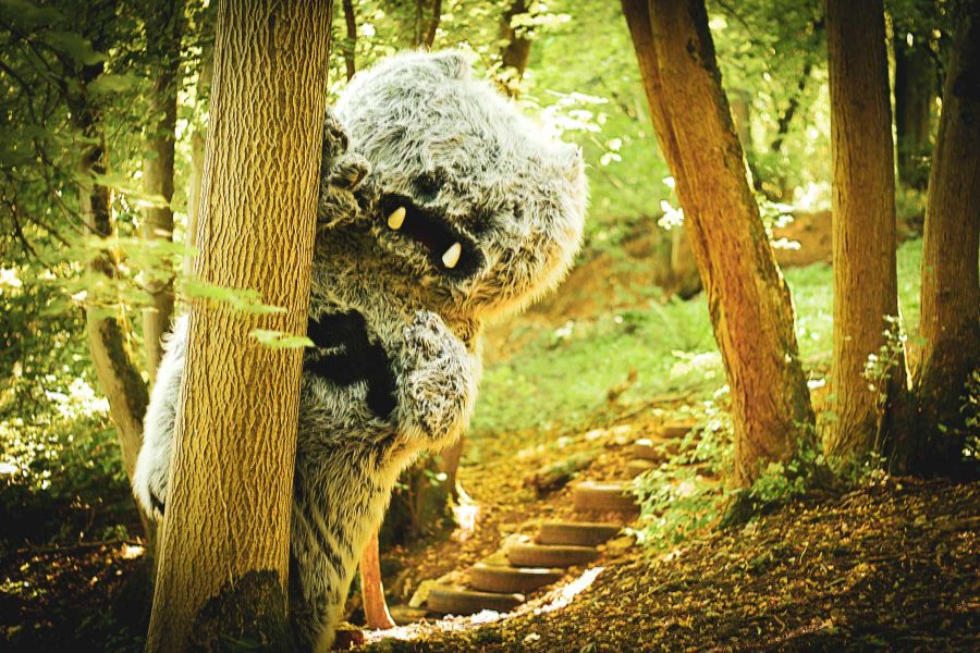 The fluffy Mendip Yeti peeks out from behind a tree in a woodland, trying to avoid being spotted by Frisbee Golfers on a Yeti Hunt - a great family holiday activity.