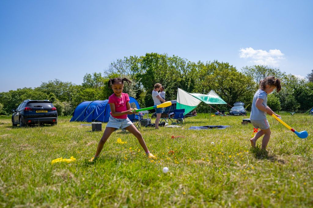 Two young girls play with a children’s golf set in the camping field. The sun is shining and the grass is green, and their mother stands in the background holding a toddler.