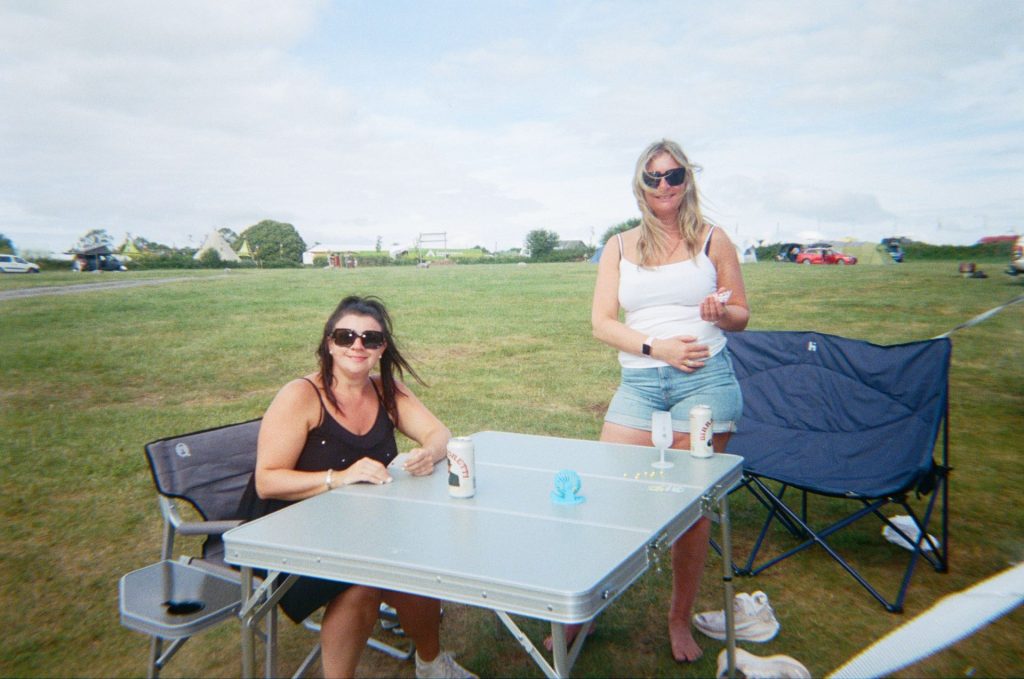 Two women are smiling and relaxing around a folding camp table with drinks.