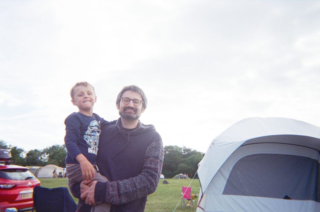 A father and son smile for the camera while standing in front of a tent. There are other cars and tents in the background, and the campsite is surrounded with mature trees.