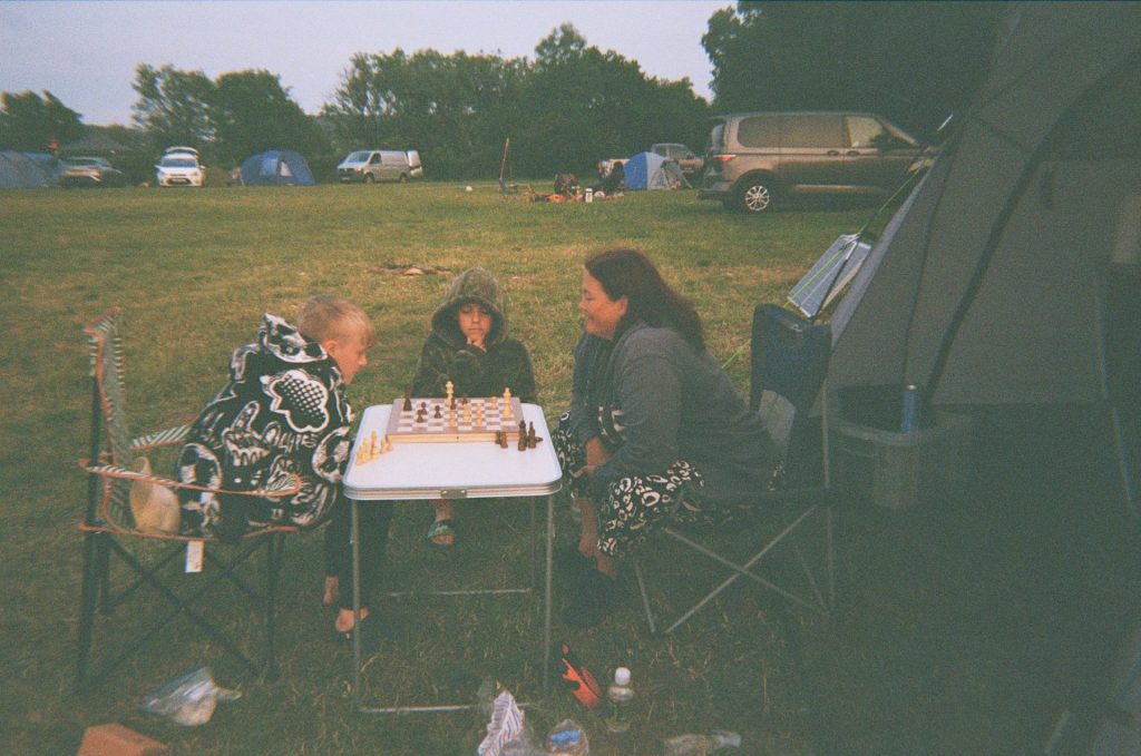 A woman and two boys at a campsite sit in folding chairs at a camping table. Two of them are playing chess while the third looks on.