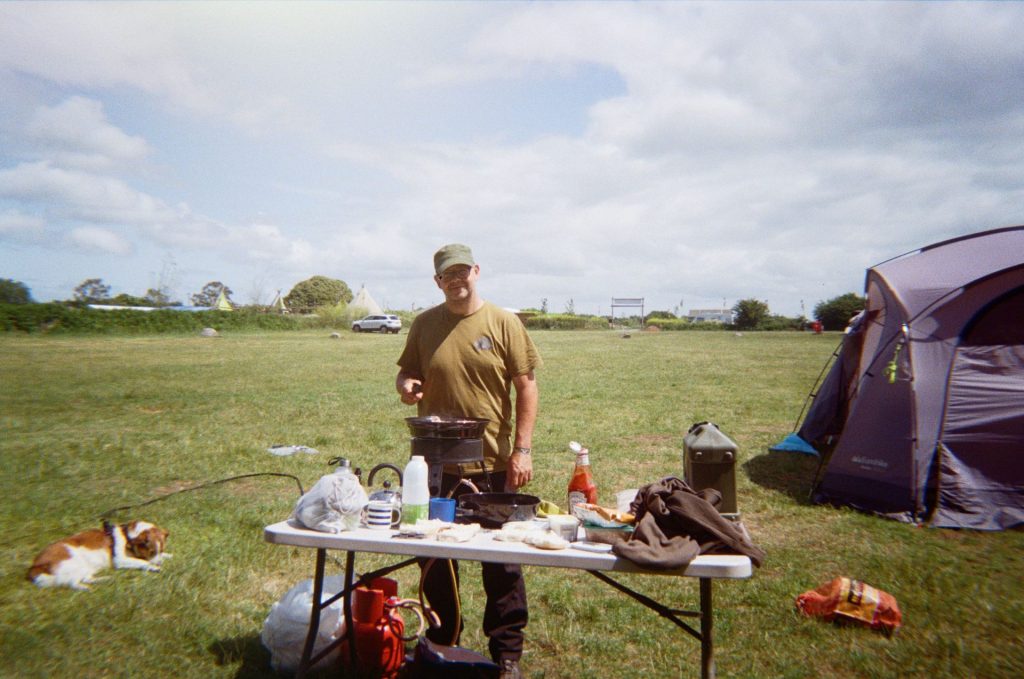 A man wearing a hat stands at a table, cooking something on a gas stove. He is in a grassy camping field; there is a tent behind him and to the left there is a small brown and white dog lying in the grass.