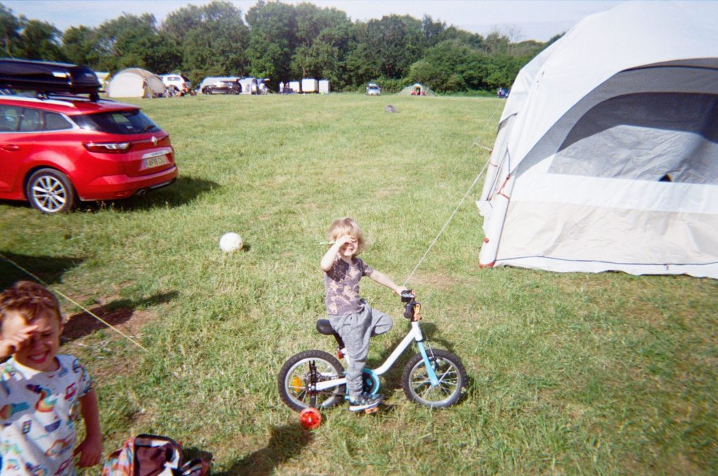 A young blonde-haired child on a bicycle smiles as he covers his eyes against the sun. Another child to the left of the image also smiles; there are camping tents and cars in the background.