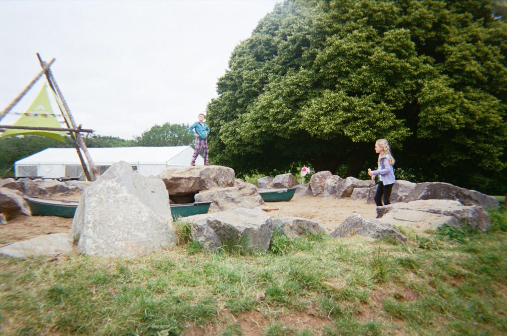 A boy and a girl explore a giant outdoor sandpit at a family adventure campsite. there is a large tree in the background and the sandpit is framed by large boulders.