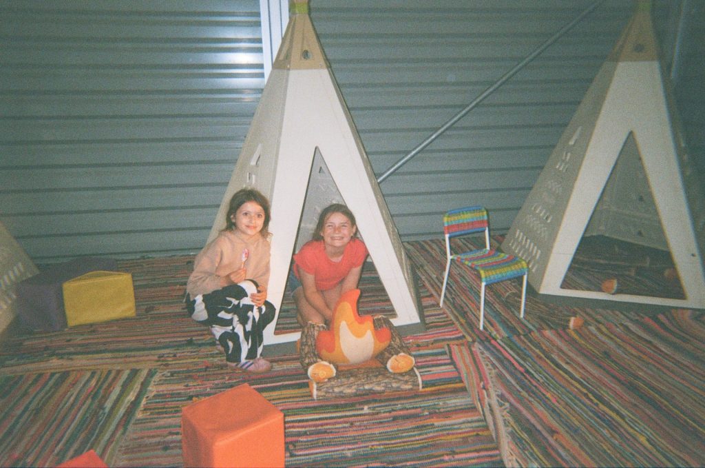 Two girls take advantage of the free activities as part of a 3 day family camping itinerary. They crouch inside a small teepee next to a pretend campfire and are smiling.