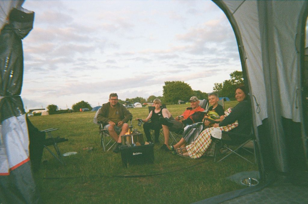 A  mixed group of adults and children sit in folding camp chairs, looking in the tent towards the viewer. The doorway of the tent frames the image; there is green grass and trees and everyone is smiling.