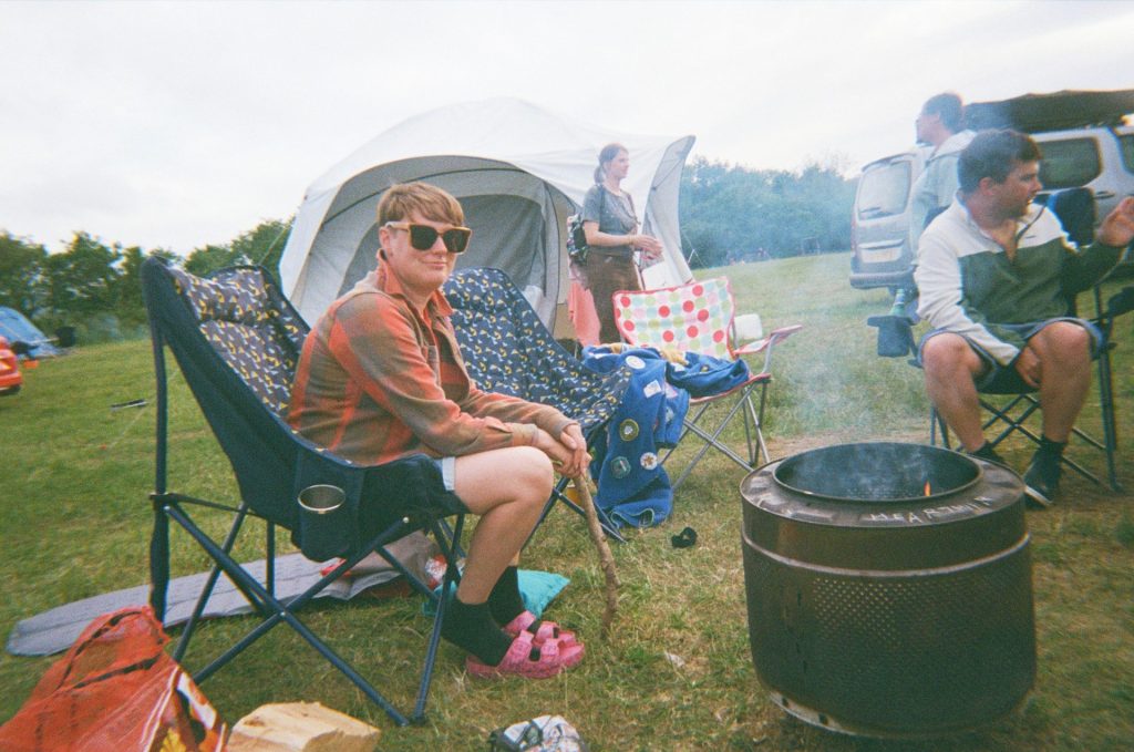 People congregate around a lit campfire at a campsite, relaxing in folding chairs or standing around and chatting.
