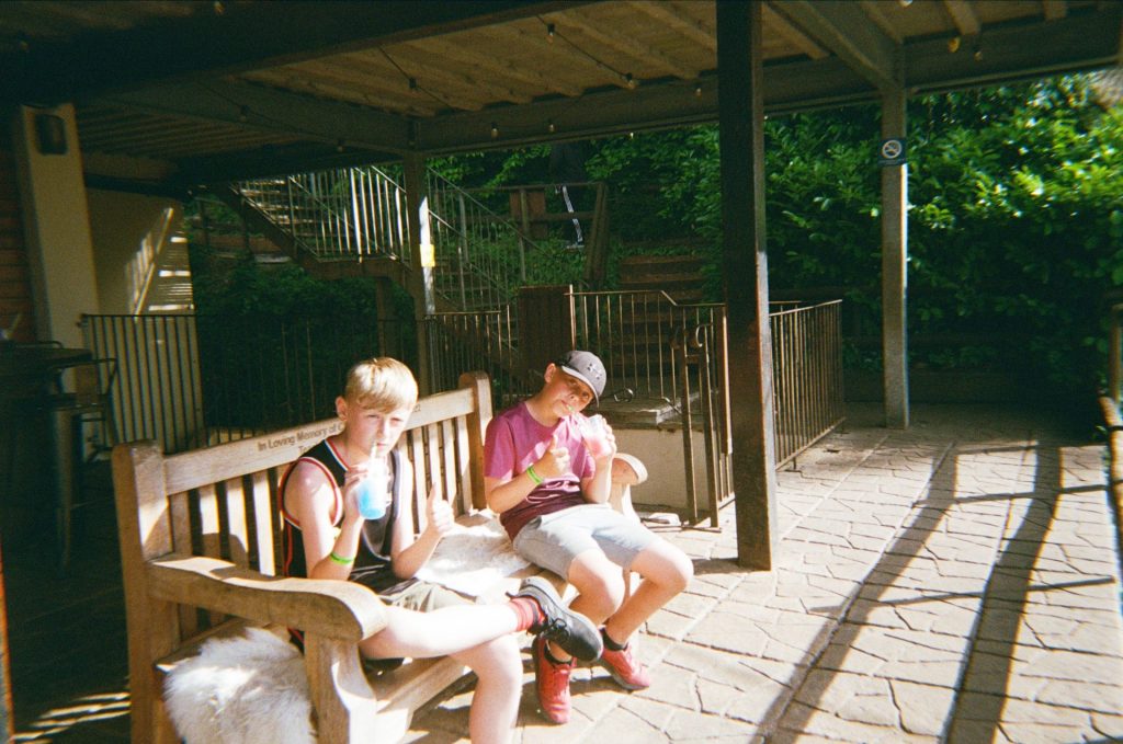 Two boys sit on a wooden bench on a patio, sipping slushies in the sunshine.