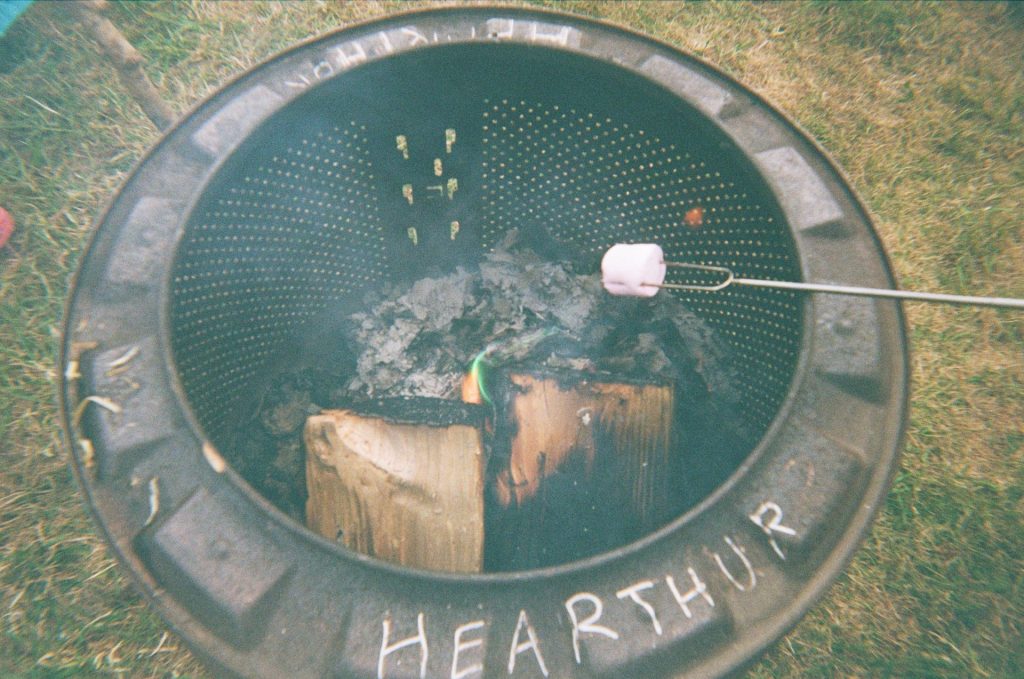 A close view of a campfire in a firepit named 'Hearthur'. Someone is holding a toasting fork with a marshmallow speared on it over the flames.