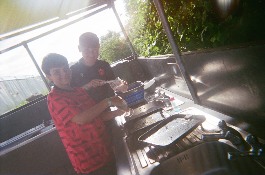 Two boys are washing dishes at the campsite's facilities. They look very happy as the sun streams in the open sides of the washing-up station.