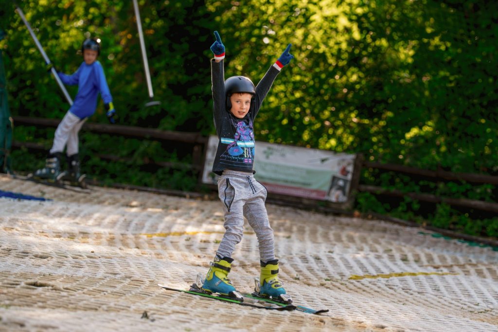 A boy triumphantly raises his arms as he skies down the all-weather dry ski slope at Mendip Activity Centre near Bristol. He is wearing a ski helmet, skis, boots and gloves.