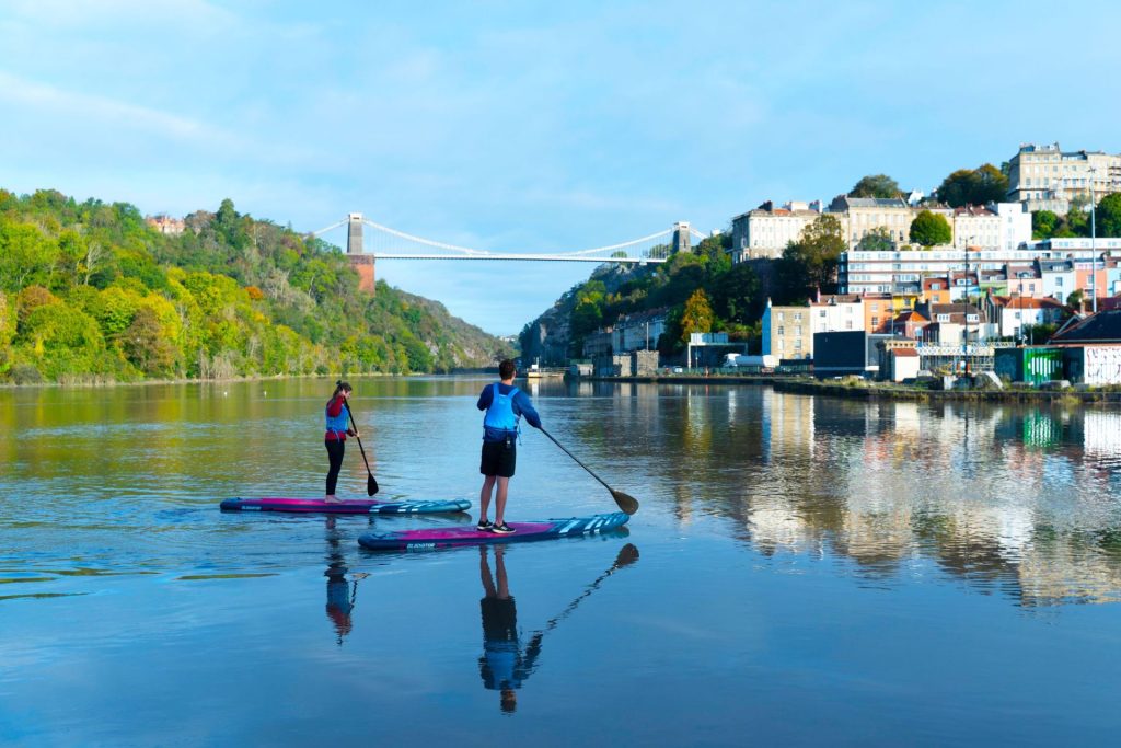 A man and a woman paddleboard near Bristol's harbourfront. They are looking towards the Clifton Suspension Bridge further up the estuary. The sun is shining and you can see some of Bristol's famous colourful terrace houses on the right.