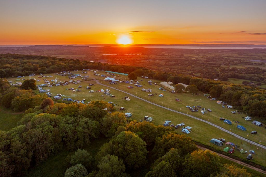 A sunset aerial view of Mendip Basecamp, a family campsite near Bristol in Somerset. A large field dotted with tents and camper vans is surrounded by woodland and far-reaching views.