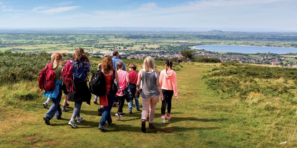 A mixed family group walks down a gently sloping, grassy footpath towards Cheddar Reservoir in the distance. There is a wide view of the Somerset Levels.