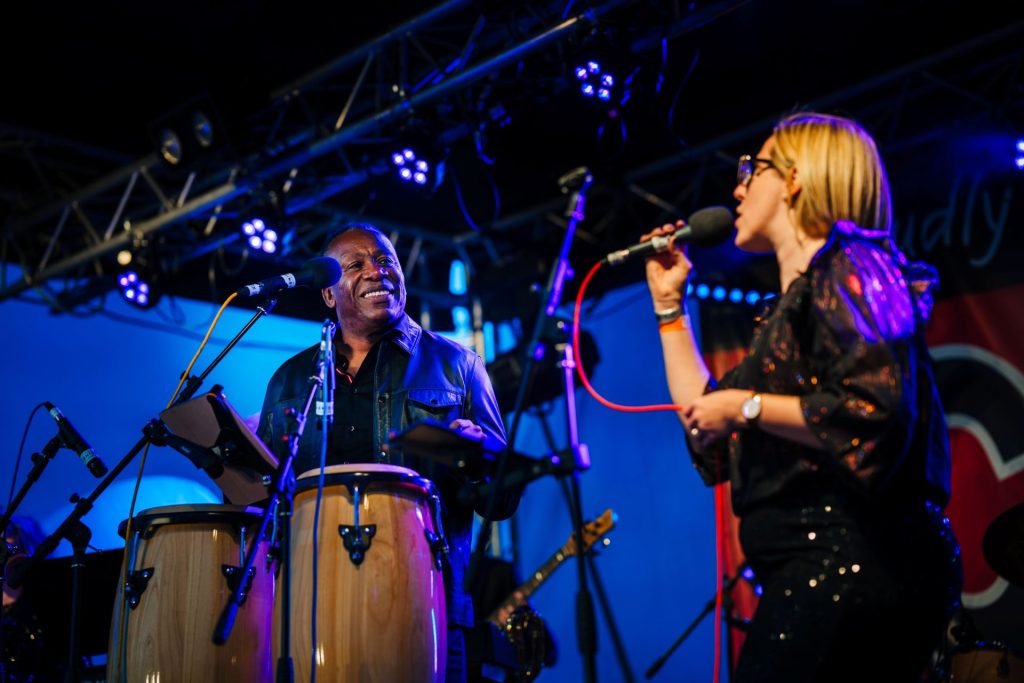 Two musicians perform live music onstage at a family festival; the man on the left plays bongo drums while looking at the woman on the right, who is singing into a handheld microphone.