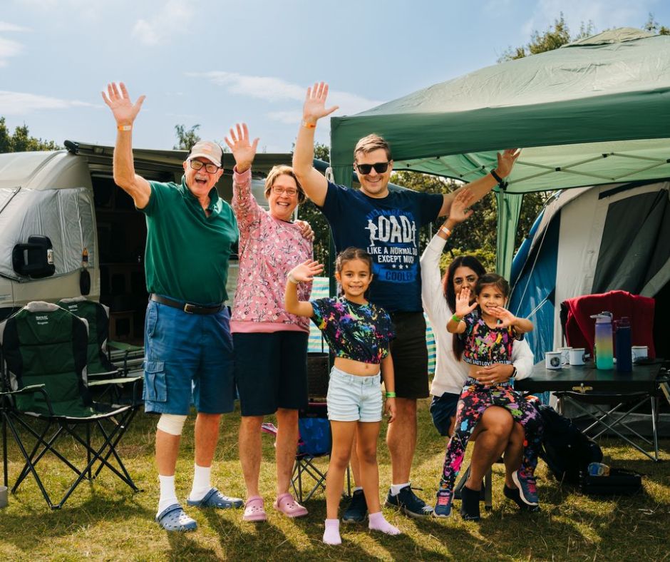 A multi-generational family group stands in front of their tent and campervan on their camping pitch, smiling and waving enthusiastically. They are surrounded by camp chairs, a table and an awning, and the sun is shining.