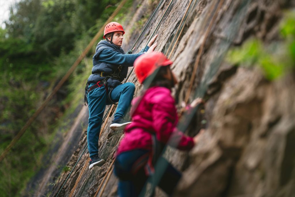 Two children are midway up a rock climbing route. They are both wearing red climbing helmets and are concentrating closely on their next move. They are outdoors, trying an adventure activity from the all-new Basecamp Bucket List.