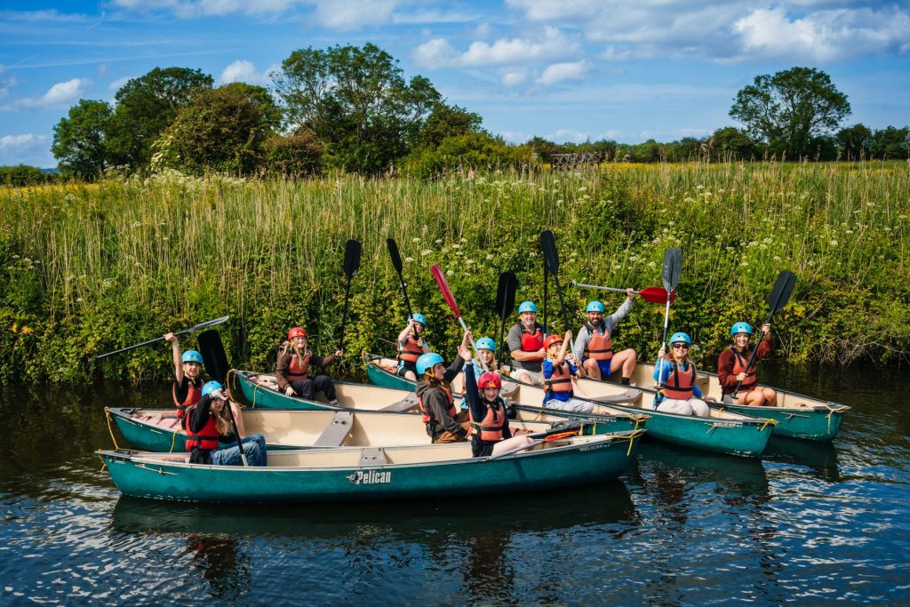 A mixed group of adults and children are sitting in canoes on the River Yeo during an adventure activity session near Cheddar Gorge. They are smiling and raising their paddles triumphantly, wearing buoyancy aids and paddling helmets.