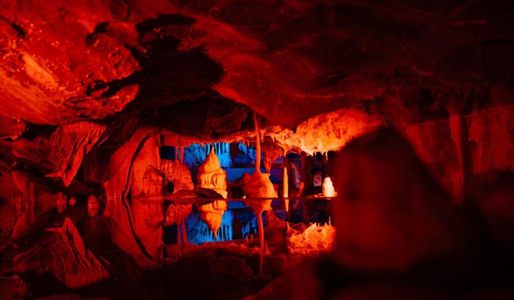 Stalactites, stalagmites and undulating rock carved by water in a Mendip Hills cave over millions of years surrounds a still, clear pool of water. It is beautifully illuminated with reddish orange light, and the rock and calcite formations are perfectly reflected in the pool. 