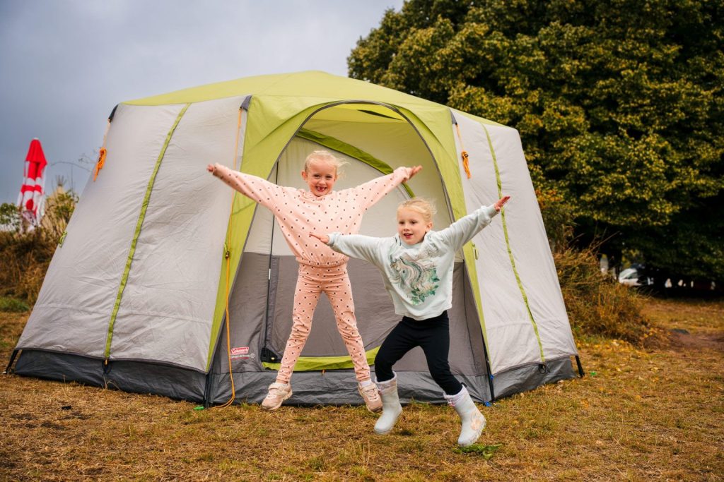 Two girls jump in the air with their arms raised, smiling. There is a green and grey tent pitched behind them, and a massive old tree further back to the left in the family campsite field.