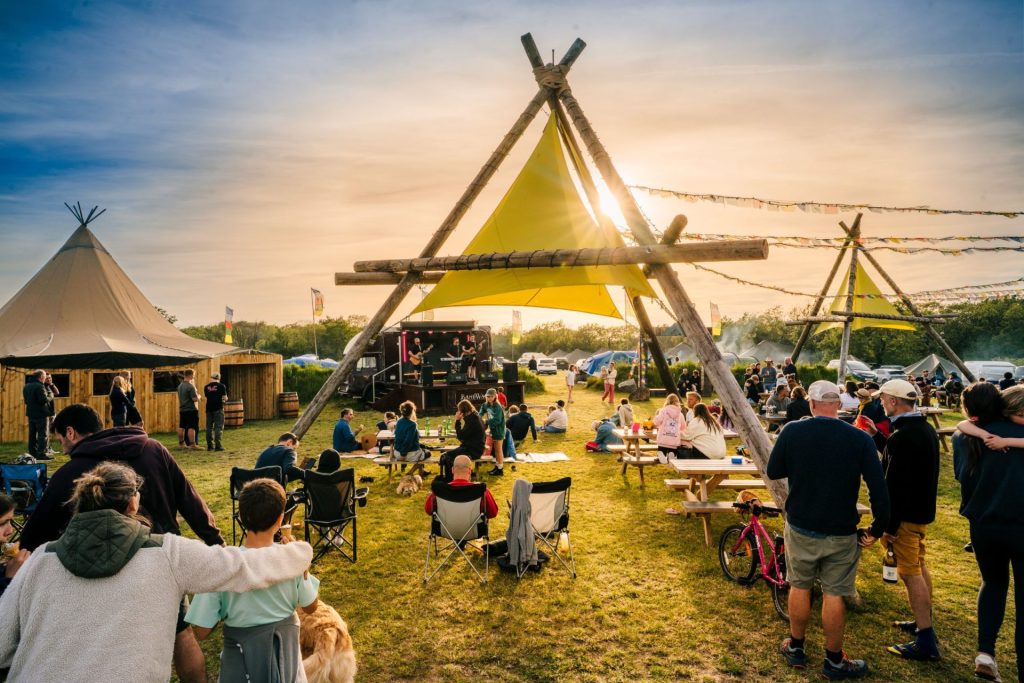 A view of the teepee area at Mendip Basecamp, decorated with flags and bunting, looking towards the setting sun. People are watching musicians play on a converted horsebox set up as a music stage. Some are relaxing on blankets on the ground, others are standing together chatting, and some are dancing to the music.