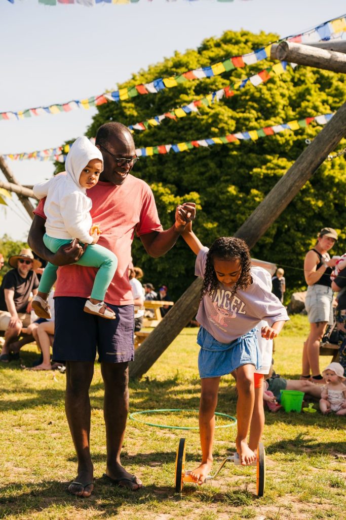 A father holds his daughter’s hand for balance as she has a go at a free circus skills activity at Basecamp Family Campsite near Bristol. She is concentrating on pedalling while her father supports her with one hand and holds another child in his other arm.