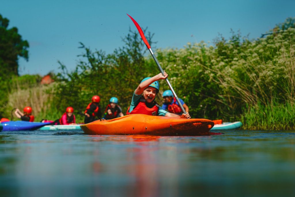 A child grins excitedly as he paddles his kayak down the Yeo River in North Somerset, on a little microadventure near Bristol. He is wearing a red buoyancy aid and blue helmet.