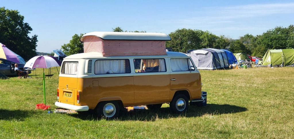 A retro orange pop-top campervan is parked in a grassy field surrounded by trees. There are tents and campervans in the background, with camping equipment like sunshades, cool boxes, camp chairs and fire pits dotted about.