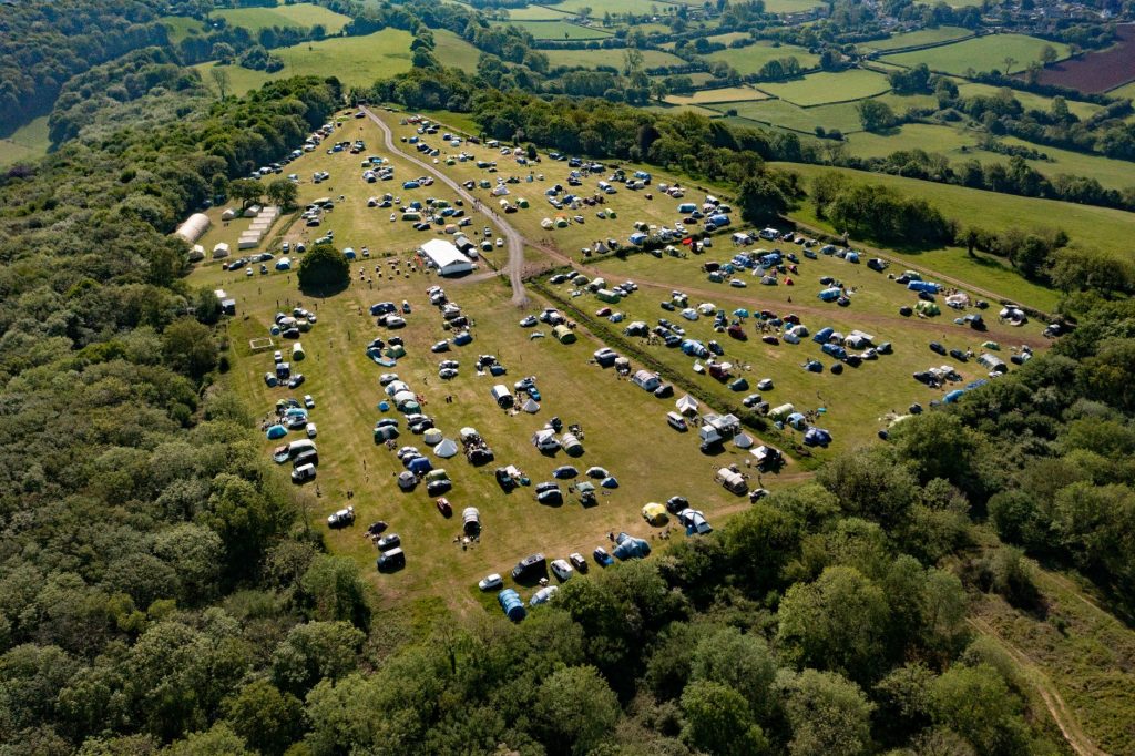 Aerial view of the Mendip Basecamp family campsite in the Mendip Hills in Somerset, a Centre Parcs alternative holiday destination. Tents and campervans are dotted around the field, which is surrounded by woodland.