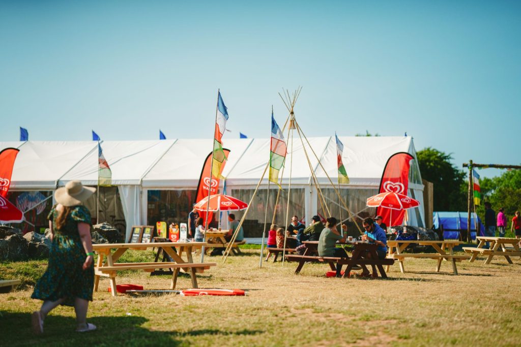 Picnic tables with red umbrellas are arranged around a large wood-pole teepee frame. A large white pavilion tent housing the cafe, camp shop and indoor play area stands in the background. Colourful flags on poles wave in the wind, giving a summery, festive feel while families walk around and sit at tables enjoying their food and drink at this Centre Parcs alternative family holiday destination.