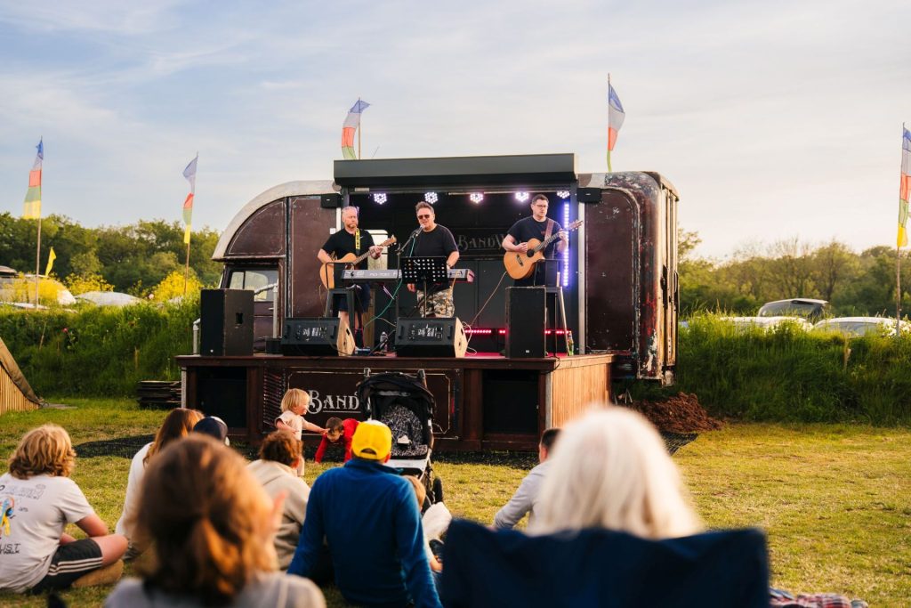 Three musicians perform on an old horsebox converted into a portable music stage at a Mendip Mini Fest at Basecamp, an alternative family holiday destination to Centre Parcs. There are colourful flags waving in the background, and the people watching the band relax in their camp chairs while children dance in front of the stage.