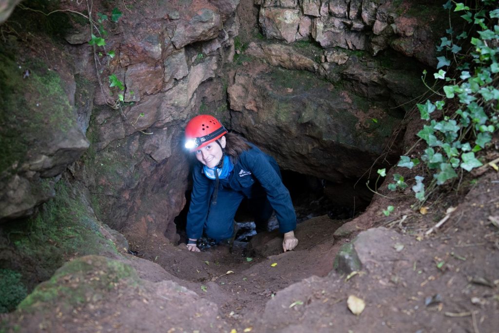 A woman doing an adventure activity in the Mendip Hills is wearing a red caving helmet and blue Mendip Activity Centre cave suit, smiling as she climbs out of a natural limestone cave.